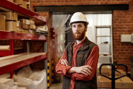 Photo for Waist up portrait of bearded worker wearing hardhat in factory warehouse and looking at camera while standing with arms crossed - Royalty Free Image