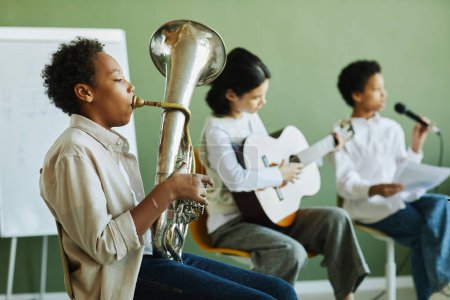 Foto de Lindo colegial afroamericano tocando la trompeta contra dos compañeros de clase interculturales tocando la guitarra y cantando - Imagen libre de derechos