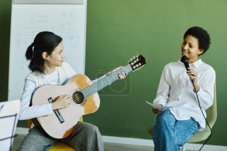 Téléchargez les photos : Deux pré-adolescents interculturels de l'école de musique interprètent une nouvelle chanson tandis que l'un d'eux chante au microphone et l'autre joue de la guitare - en image libre de droit