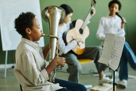 Photo for Side view of pre-teen schoolboy blowing wind instrument against two intercultural classmates playing guitar and singing in microphone - Royalty Free Image