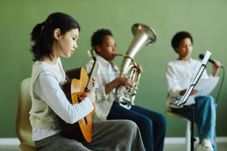 Téléchargez les photos : Mignon asiatique écolière jouer de la guitare acoustique et regarder des notes sur le stand de musique tout en étant assis contre deux camarades de classe à la leçon - en image libre de droit