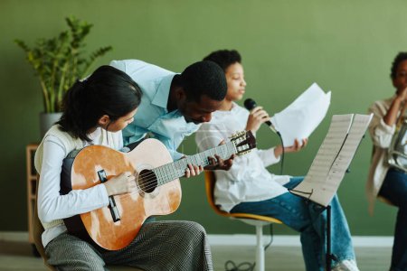 Photo for Young African American male teacher of music bending over pre-teen Asian schoolgirl with acoustic guitar while consulting her - Royalty Free Image
