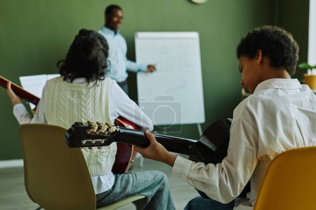 Téléchargez les photos : Vue arrière de deux jeunes apprenants de l'école de musique jouant des instruments à cordes devant un professeur debout près d'un tableau blanc - en image libre de droit