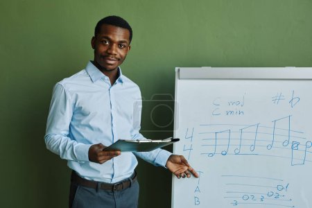Téléchargez les photos : Jeune professeur de musique à succès avec un document pointant vers le tableau blanc avec des notes musicales pendant l'explication et regardant la caméra pendant la leçon - en image libre de droit