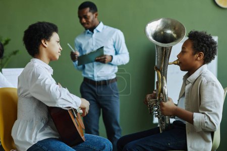 Photo for Two youthful schoolkids playing trumpet and acoustic guitar at lesson while sitting in front of each other with their teacher on background - Royalty Free Image