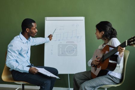 Photo for Side view of confident teacher of music explaining musical notes to student with acoustic guitar while pointing at whiteboard - Royalty Free Image