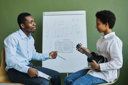 Téléchargez les photos : Jeune professeur afro-américain confiant répondant aux questions de l'écolière à la leçon avec ukulele tout en étant assis au tableau blanc - en image libre de droit