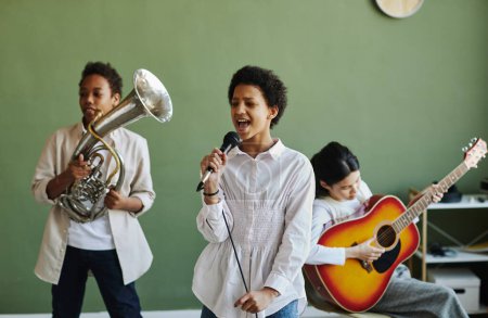 Téléchargez les photos : Heureuse écolière jeune avec microphone chantant avec deux camarades de classe jouant de la trompette et de la guitare acoustique à la leçon - en image libre de droit