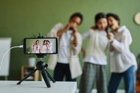 Téléchargez les photos : Écran du smartphone sur trépied avec trois écoliers chantant ensemble tout en enregistrant une vidéo ou en faisant de la diffusion en direct - en image libre de droit