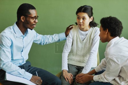 Photo for Young African American male psychologist supporting Asian schoolgirl and giving her advice at session while keeping hand on her shoulder - Royalty Free Image