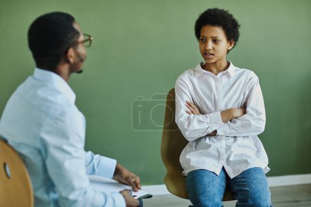 Photo for Youthful anxious schoolgirl describing her problem to counselor at psychological session while sitting with her arms crossed by chest - Royalty Free Image