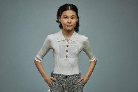 Téléchargez les photos : Jeune écolière d'origine asiatique portant un pantalon gris et un pantalon blanc gardant les mains dans les poches et regardant la caméra - en image libre de droit