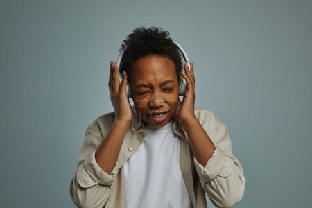 Foto de Lindo chico de la generación Alpha tocando auriculares en su cabeza mientras mantiene los ojos cerrados y disfruta de su música favorita - Imagen libre de derechos