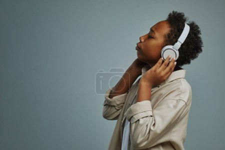 Téléchargez les photos : Adorable garçon pré-adolescent en chemise décontractée profitant de sa musique préférée dans les écouteurs tout en posant pendant la séance photo en studio - en image libre de droit