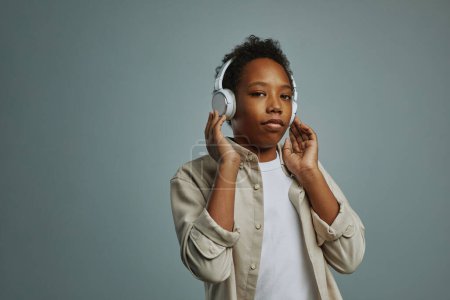 Photo for Youthful schoolboy touching white headphones while listening to his favorite music and looking at camera over grey background - Royalty Free Image