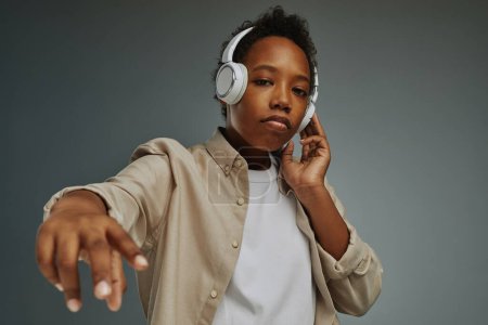 Photo for Cute African American boy in headphones looking at camera while standing in isolation on grey background and enjoying rap music - Royalty Free Image