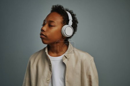 Photo for Cute serene boy enjoying relaxation music in white headphones while keeping eyes closed and posing for camera on grey background - Royalty Free Image