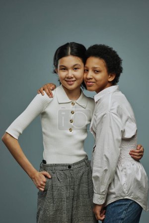 Photo for Two adorable intercultural schoolgirls in white casual tanktops embracing one another and looking at camera while posing over grey background - Royalty Free Image