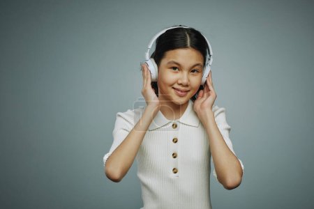 Photo for Smiling youthful Asian girl touching headphones while listening to her favorite music and performer against grey background in studio - Royalty Free Image