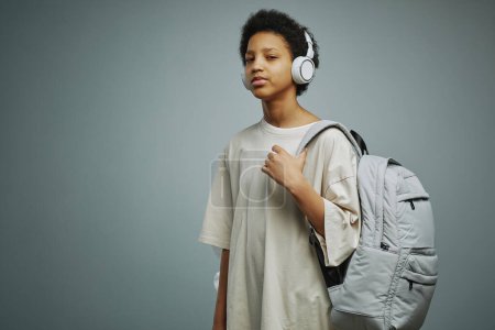 Téléchargez les photos : Écolière américaine AFricane mignonne avec sac à dos sur l'épaule et écouteurs sur la tête écoutant de la musique et regardant la caméra - en image libre de droit