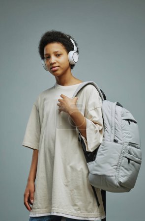 Téléchargez les photos : Écolière mignonne en t-shirt blanc long et headphnes tenant le sac à dos sur l'épaule et vous regardant tout en posant devant la caméra - en image libre de droit