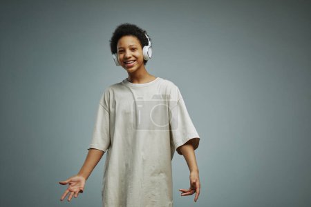Téléchargez les photos : Joyeux afro-américaine écolière en long t-shirt blanc riant et regardant la caméra tout en appréciant la musique énergique dans les écouteurs - en image libre de droit