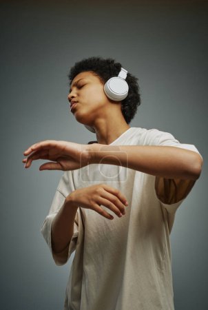 Téléchargez les photos : Jeune fille afro-américaine en t-shirt long profitant de sa musique préférée dans les écouteurs tout en gardant les mains devant elle pendant la danse - en image libre de droit