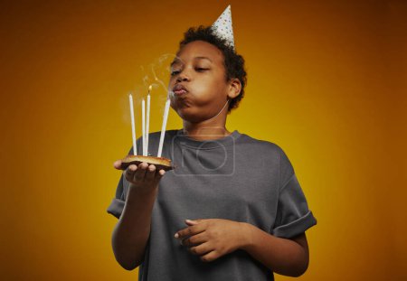 Photo for Cute African American youngster in birthday cap and grey t-shirt blowing burning candles while making wish for his birthday in studio - Royalty Free Image