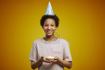 Téléchargez les photos : Joyeux afro-américaine pré-adolescente avec morceau de gâteau d'anniversaire avec plusieurs bougies allumées debout devant la caméra en isolement - en image libre de droit
