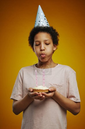 Téléchargez les photos : Jeune fille dans le chapeau d'anniversaire soufflant des bougies sur petit gâteau fait maison tout en se tenant devant la caméra et faire le vœu à la fête - en image libre de droit