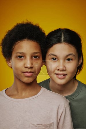 Photo for Portrait of two adolescent friendly girls in t-shirts standing in front of camera against bright yellow background and looking at you - Royalty Free Image