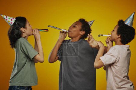 Photo for Group of three intercultural pre-teen children in t-shirts and birthday caps blowing whistles while having fun and enjoying party - Royalty Free Image