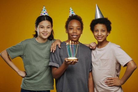 Photo for Three youthful multicultural friends in birthday caps and casualwear standing in front of camera while boy holding cake with candles - Royalty Free Image
