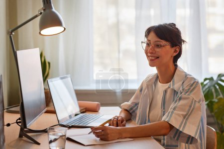Téléchargez les photos : Portrait d'une jeune femme souriante profitant d'un cours d'éducation en ligne ou d'une réunion de travail au bureau à domicile confortable avec des lumières tamisées - en image libre de droit