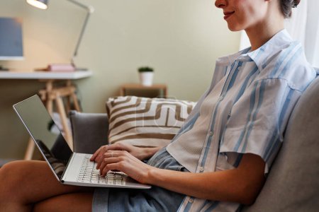 Téléchargez les photos : Portrait recadré d'une jeune femme souriante utilisant un ordinateur portable assis sur un canapé à la maison et travaillant - en image libre de droit