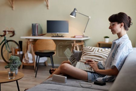 Téléchargez les photos : Portrait de vue latérale d'une jeune femme souriante utilisant un ordinateur portable tout en se relaxant sur un canapé dans un intérieur confortable, espace de copie - en image libre de droit