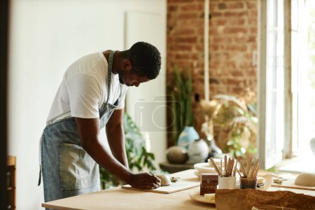 Téléchargez les photos : Portrait latéral de jeune homme afro-américain créant un beau bol fait à la main dans un studio de poterie confortable, espace de copie - en image libre de droit