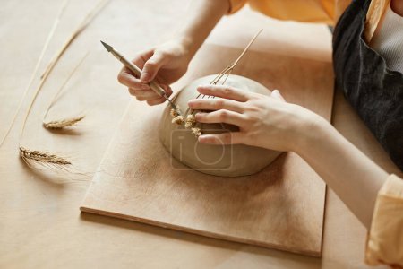 Téléchargez les photos : Gros plan de l'artisan féminin décorant à la main des céramiques avec une empreinte végétale à l'aide d'une fleur immortelle, espace de copie - en image libre de droit