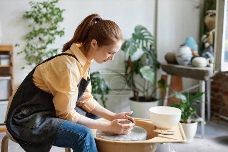 Téléchargez les photos : Portrait latéral de jeune femme utilisant la roue de poterie et la création de céramiques faites à la main dans un studio confortable - en image libre de droit