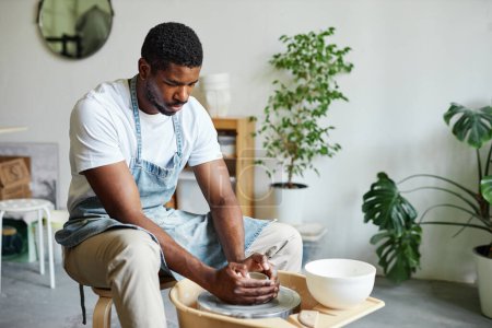Téléchargez les photos : Portrait de jeune homme noir utilisant la roue de poterie et la création de céramique à la main dans un studio confortable avec des plantes vertes - en image libre de droit