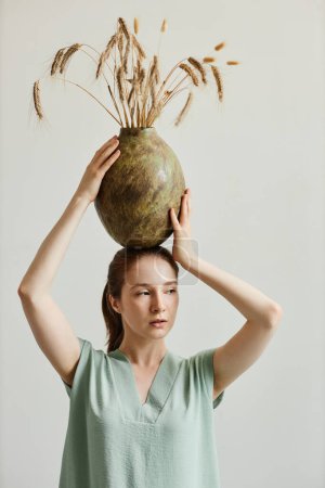 Photo for Minimal portrait of elegant young woman balancing handmade ceramic vase on head while standing against white background, raw beauty - Royalty Free Image