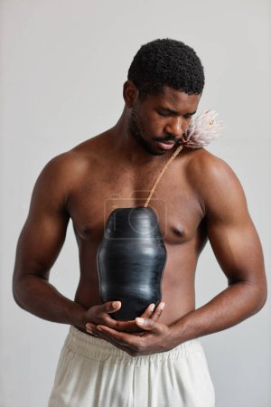 Photo for Minimal portrait of sensual black man shirtless and holding tan ceramic vase with exotic flower - Royalty Free Image