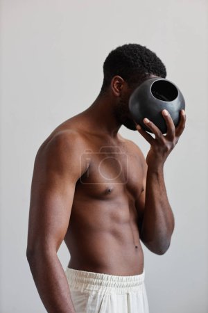 Photo for Minimal concept portrait of shirtless black man holding tan ceramic vase and hiding face, natural shapes in design - Royalty Free Image