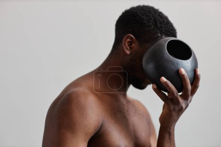 Téléchargez les photos : Minimal concept shot of shirtless black man holding rownd tan vase and hide face, formes naturelles dans la conception wabi sabi - en image libre de droit