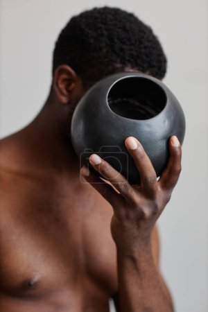 Photo for Vertical shot of shirtless black man holding round tan vase and hiding face, nature in design - Royalty Free Image