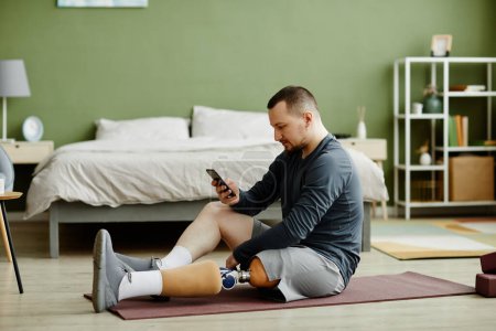 Foto de Retrato de vista lateral de un hombre adulto con una pierna protésica sentado en una esterilla de yoga en casa y usando un teléfono inteligente - Imagen libre de derechos