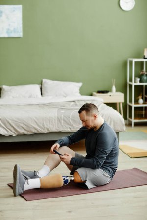 Photo for Vertical side view portrait of adult man with prosthetic leg sitting on yoga mat at home and using smartphone - Royalty Free Image
