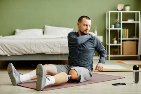 Foto de Retrato completo del hombre con la pierna protésica haciendo ejercicios de estiramiento en la esterilla de yoga en casa - Imagen libre de derechos