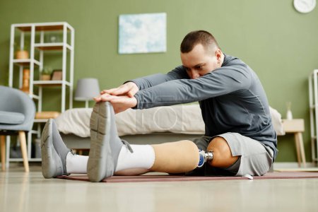 Téléchargez les photos : Portrait complet de l'homme avec la jambe prothétique faire des exercices d'étirement à la maison et atteindre les orteils - en image libre de droit