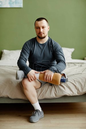 Téléchargez les photos : Portrait vertical de l'homme tenant une jambe prothétique assis sur le lit et regardant la caméra - en image libre de droit